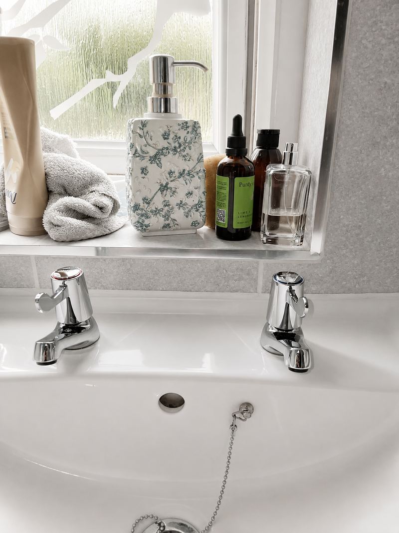 A pair of chrome pillar taps on a white bathroom basin, with toiletries on the windowsill behind