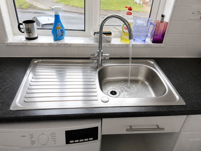 Stainless steel kitchen sink with a chrome swan-neck mixer running, on a black worktop beside a washing machine