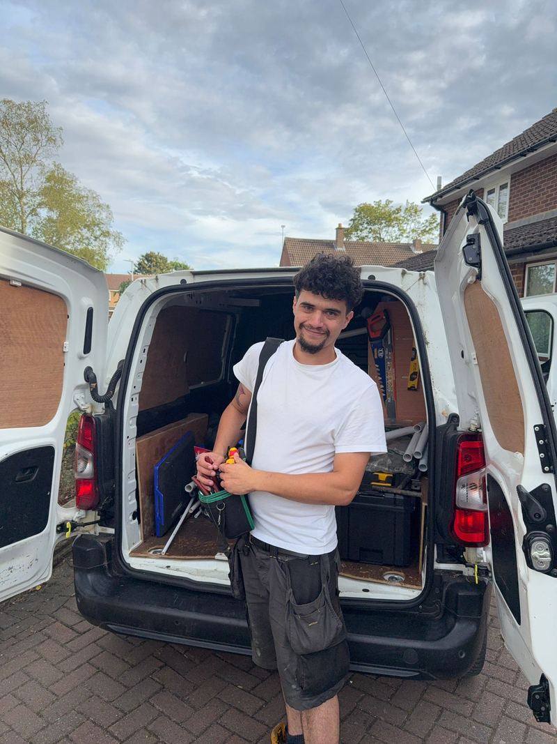 Tony from AGL Plumbing Services at the open back doors of his white work van, holding a pipe wrench, with tools and pipe stacked inside