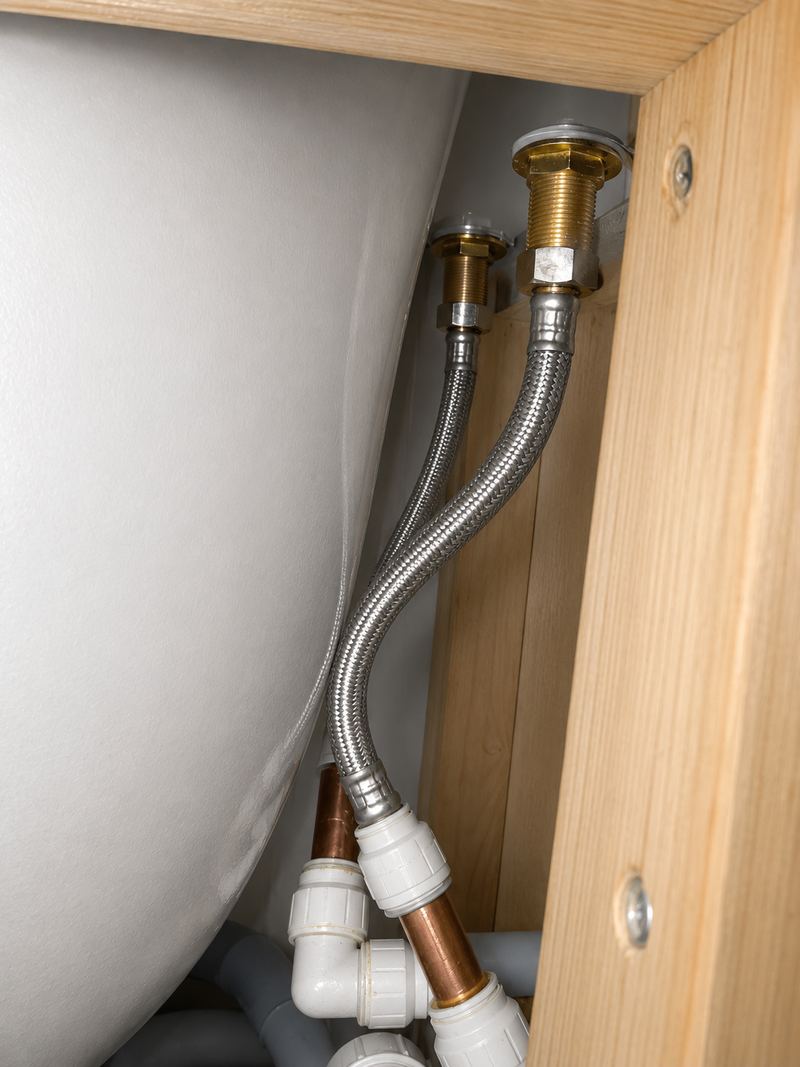 Two braided flexible tap connectors running from brass tap tails down to white push-fit and copper pipework inside an oak sink cupboard
