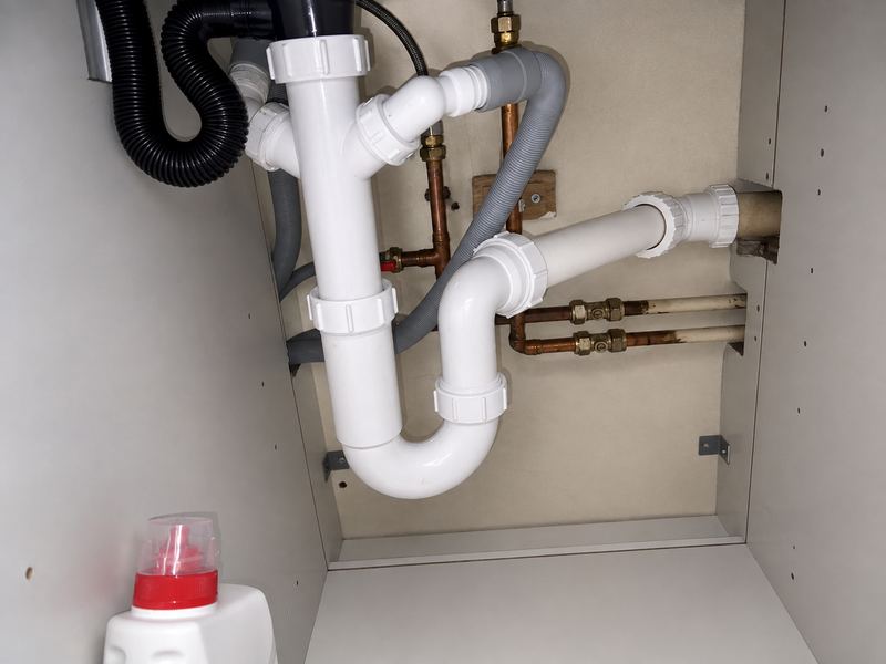 White plastic waste pipework and a P-trap under a kitchen sink, with copper feed pipes, isolation valves and an appliance drain hose